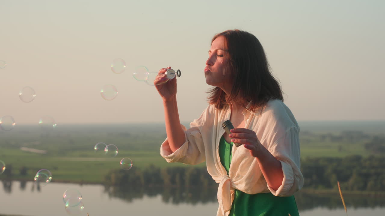 Fair skinned woman in white blouse and green dress on cliff edge overlooking river valley at sunset, gently blowing iridescent soap bubbles drifting through tall grass under pastel evening sky