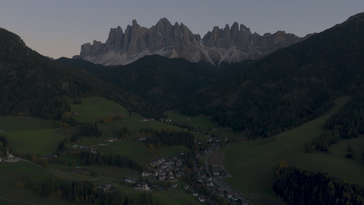 Vista aerea cinematografica delle Dolomiti italiane all'alba, aspre cime rocciose sopra rigogliose colline alpine e chalet sparsi, sereno paesaggio naturale di montagna