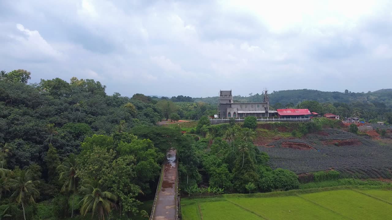 Aerial shot of a narrow aqueduct over a valley of paddy fields, leading toward a large, imposing Christian church under construction on a tropical hillside with pineapple plantation