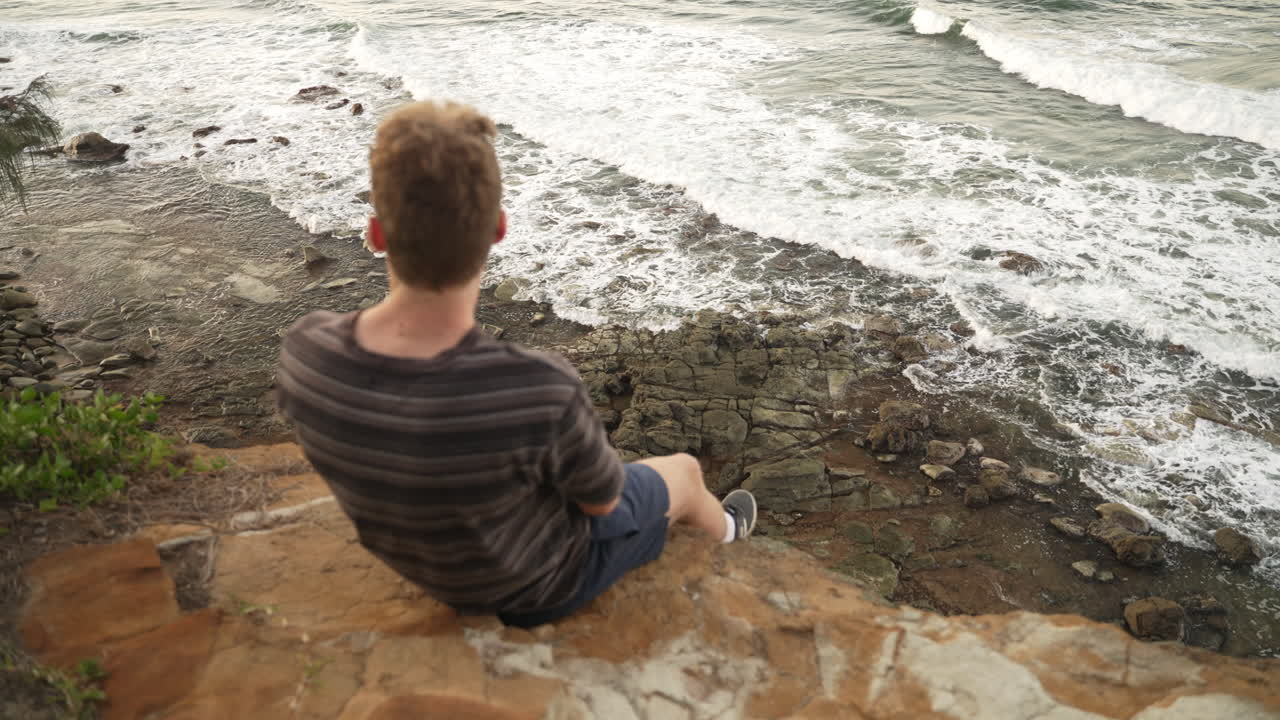 바위가 많은 해안이 내려다 보이는 바위 절벽에 앉아 있는 한 젊은 남성, 4k