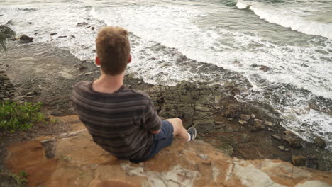 Single-Young-Male-Sitting-On-Rocky-Cliff-Overhang-Overlooking-Rocky-Shore-And-Crashing-Ocean-Waves,-4K