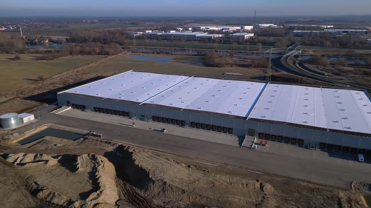 Nuovo grande edificio del centro logistico in costruzione con una vasta area di stoccaggio, banchine di carico e terreno vuoto circostante. Favoloso panorama aereo orbitale con drone