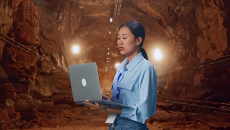 Side View Of Asian Female Professional Worker In Underground Mine Tunnel, Observes By Looking Up Before She Come To Concentrating On The Laptop And Keep On Typing