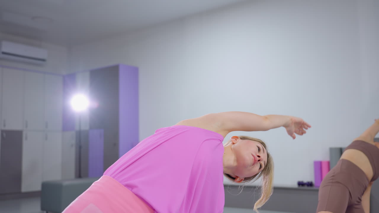 mujeres que participan en un entrenamiento de estiramiento enfocado en el gimnasio, extendiendo los brazos mientras mantienen el equilibrio, el fondo presenta equipos de gimnasio y un instructor que ayuda con la postura correcta