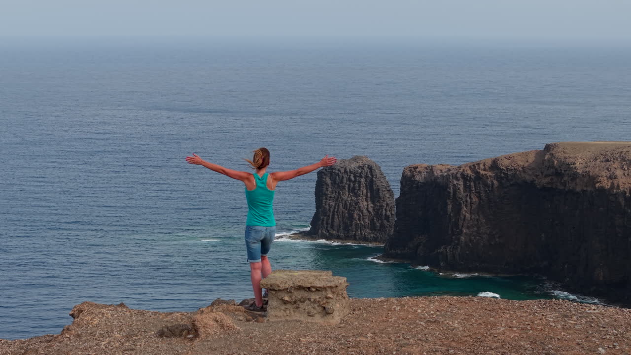 Female traveler standing on rocky volcanic cliff, overlooking expansive Atlantic Ocean coastline with panoramic Gran Canaria landscape under bright sunlight