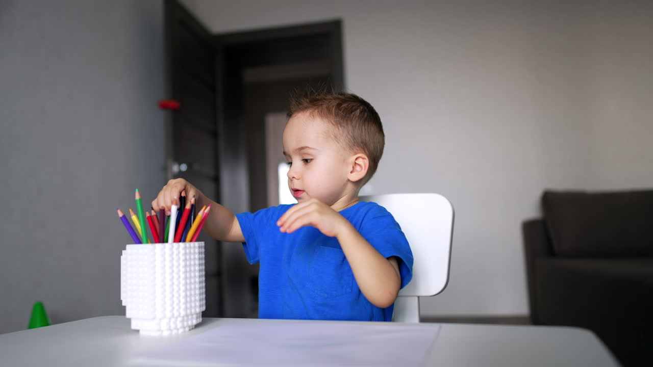 Caucasian child sitting at desk in his room chooses a pencil from a cup. Baby boy learning colors and doing art.