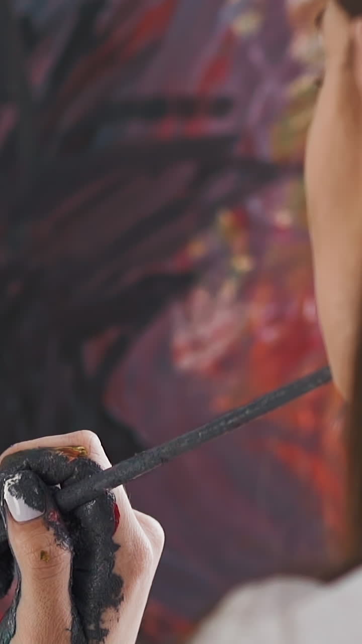 Brunette lady artist holds brush with dirty hand looking at canvas with abstract drawing in contemporary studio extreme close back view slow motion