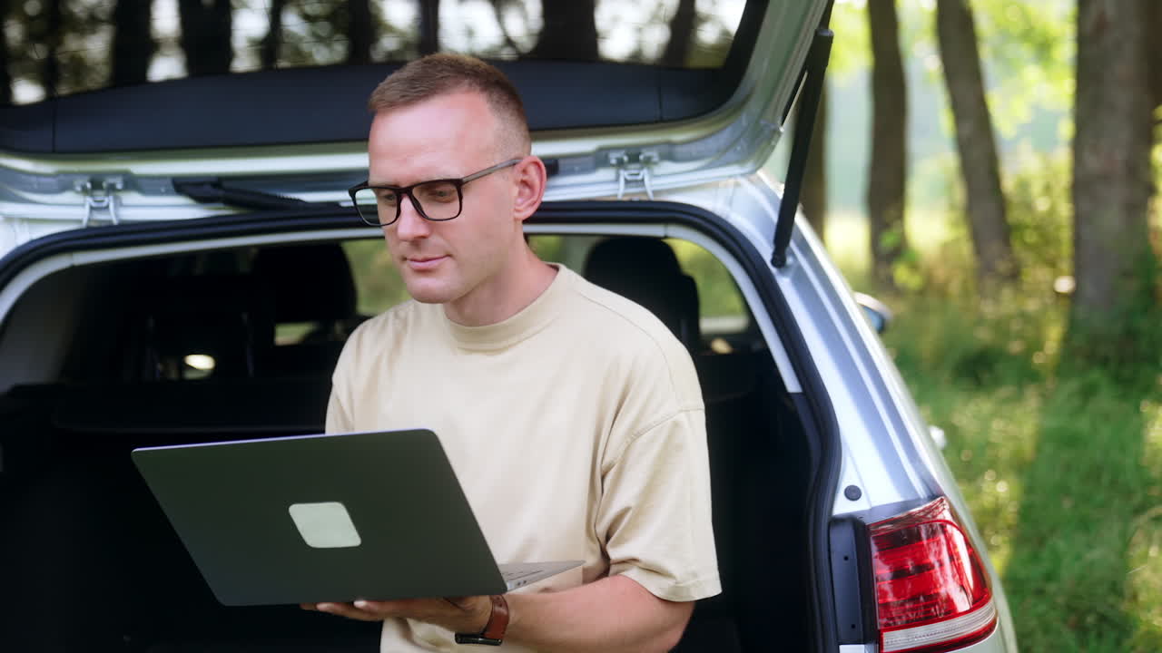 Mid-aged freelancer focused on laptop. Man works sitting in car trunk in the forest.