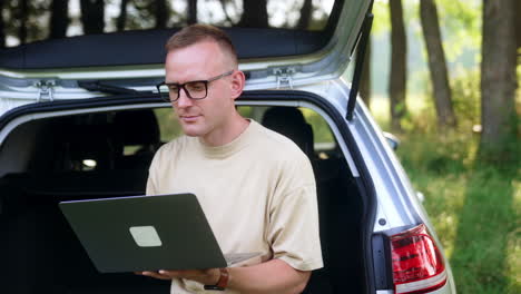 mid-aged freelancer focused on laptop. man works sitting in car trunk in the forest.
