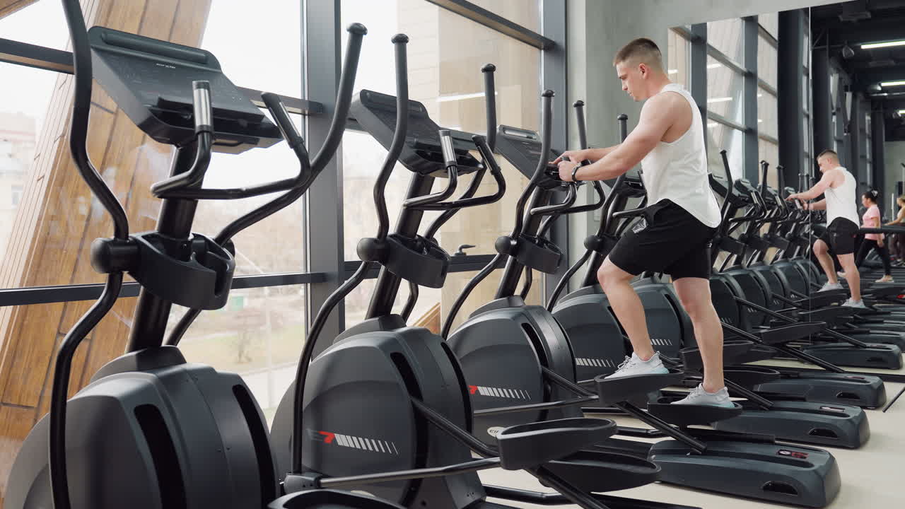 Fitness enthusiast works out on elliptical trainer in bright modern gym next to mirror reflecting him and others during active cardio exercise session beneath large windows and hanging straps