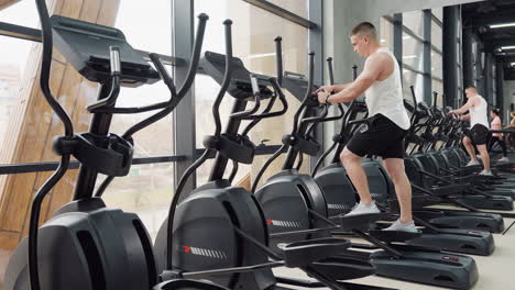 fitness enthusiast works out on elliptical trainer in bright modern gym next to mirror reflecting him and others during active cardio exercise session beneath large windows and hanging straps
