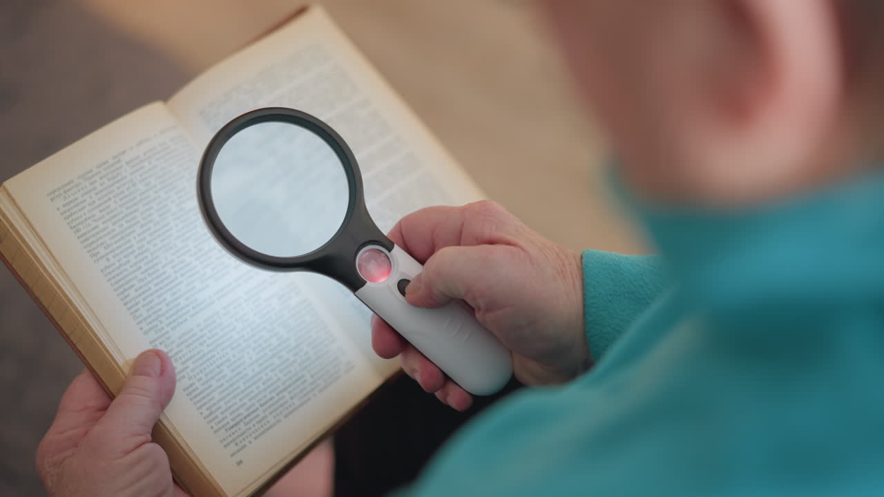 Close-up shot of elderly person using magnifying glass on book, focusing on small print. Hand is holding magnifier with light on while reading. Soft focus on blurred book background