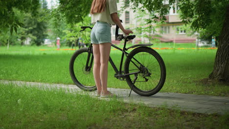 woman in pink sneakers and jean shorts approaches black bicycle on paved path, placing one glove on seat while wearing other glove, background includes people walking in distance