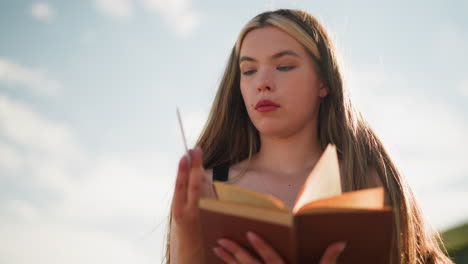 close up of thoughtful young woman in black flipping through book under bright sky, she finds a piece of paper within pages and gazes at it contemplatively, as sunlight gently illuminates her face