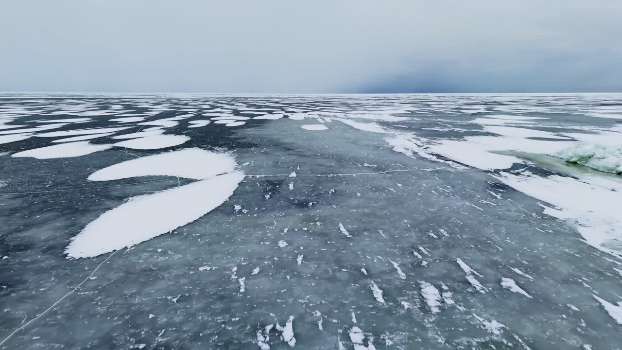 Inquadratura cinematografica di ambientazione di un vasto mare ghiacciato con ghiaccio scuro crepato e chiazze di neve sparse, ripreso in basso con drone sotto un cielo invernale coperto che sfuma nella nebbia