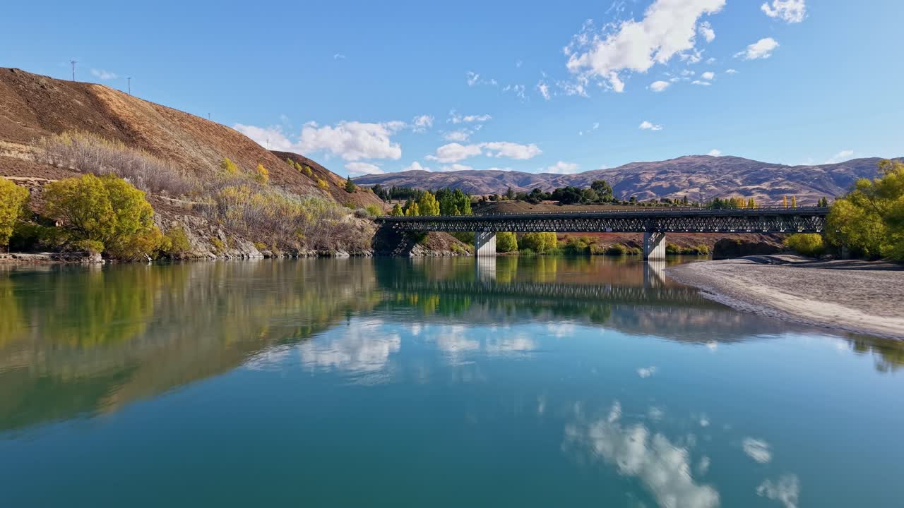 Drone glides forward above calm turquoise water toward bridge with cars crossing