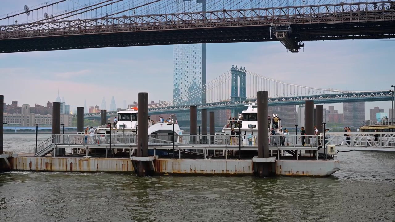 New York, USA, 1 maggio 2026: Terminal dei traghetti sull'East River vicino al Ponte di Brooklyn a New York. Barche passeggeri riposano al molo galleggiante mentre un ponte sospeso attraversa il fiume sotto un cielo coperto.