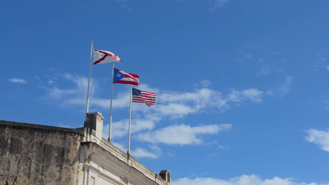 Bandiere americana e portoricana che sventolano nella brezza sulle storiche mura in pietra del forte a Vecchia San Juan, Porto Rico, sotto un cielo blu