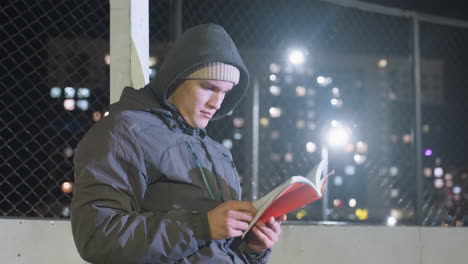 athlete in hoodie and beanie leaning against metallic goalpost outdoors at night, thoughtfully reading book under urban city lights, peaceful moment of reflection, illuminated by streetlights