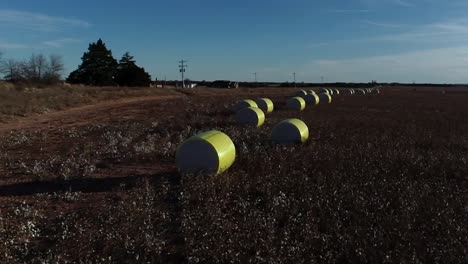 Fast-paced,-sweeping-Drone-Aerial-shot-of-a-Midwestern-cotton-farm-with-fresh-bales-of-harvested-cotton-wrapped-in-bright-yellow-material-against-a-blue-open-sky