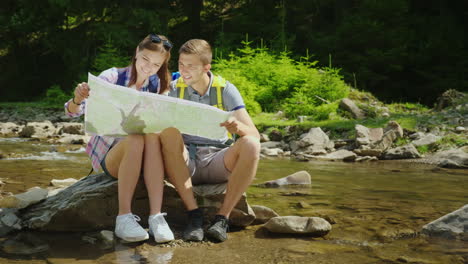 a young couple of tourists are studying together a map they sit in a picturesque place near a mounta