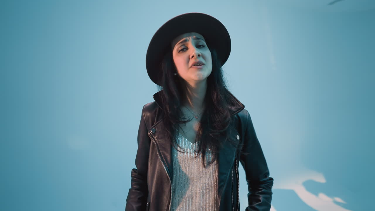 una mujer con una chaqueta de cuero negra, pantalones y sombrero canta con pasión, moviendo su cuerpo y manos expresivamente contra un fondo azul. ella actúa sin un micrófono