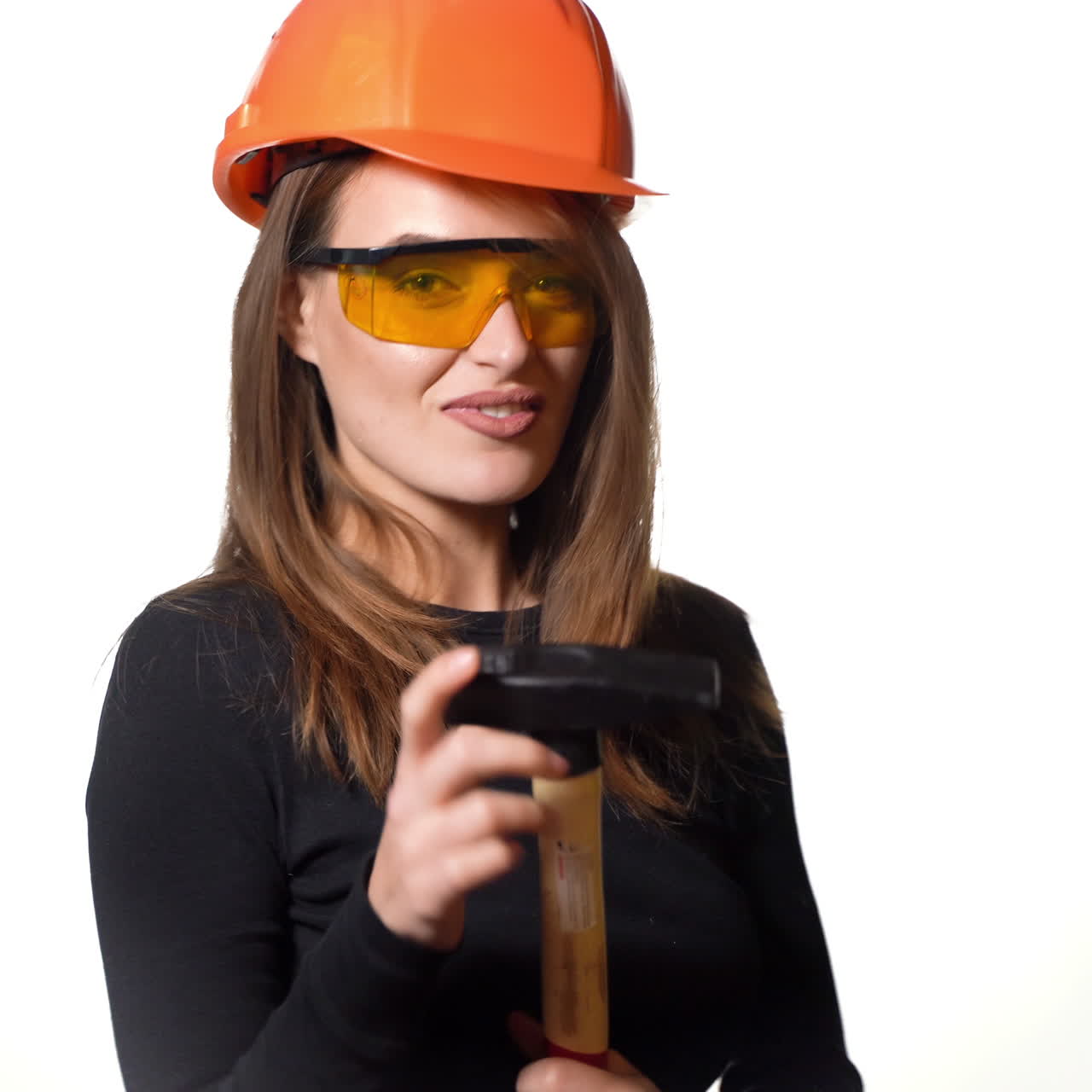 A beauty with long brown hair in an orange helmet and glasses is holding a hammer in her hand and posing with it on a white background in the studio.