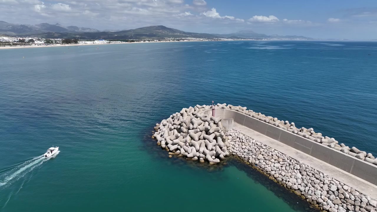 Vista aerea di un piccolo motoscafo che lascia un porto mediterraneo protetto nel Marocco settentrionale, passando tra i frangiflutti e dirigendosi verso il mare aperto e blu con una costa montuosa in lontananza