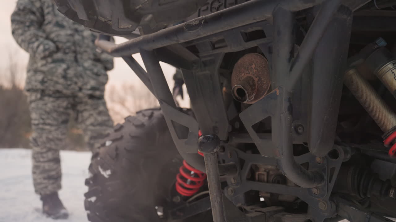 Atv rear inspection in snowy field. Camouflageclad rider checks spare tire and rear suspension on rugged quad during golden hour, steam rising from exhaust, red coil shocks visible, focused prep