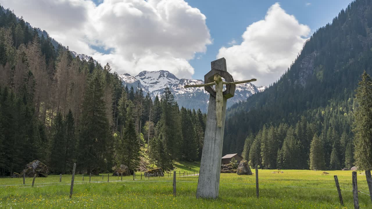 Time-lapse in front of a Jesus cross in the Swiss Alps in a valley in spring. snow-capped mountains in the background. The natural landscape blooms between the forests. clouds move in sky