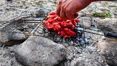 Male-hand-is-laying-meat-on-a-grill-to-bbq-it-on-the-beach-in-the-sand,-closeup-in-slow-motion