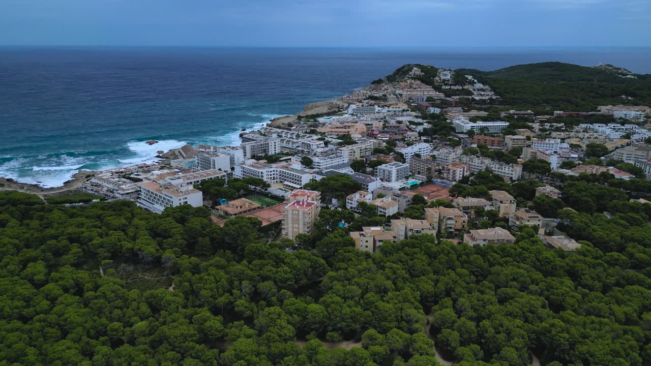 Features the wild, forested coast of Cala Agulla alongside the dense buildings, hotels, and apartments of the nearby tourist resort. Mediterranean travel and lifestyle