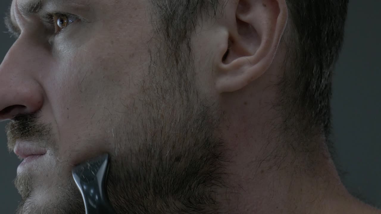 Caucasian male shaving his beard at the cheek with battery razor to shape the outline
