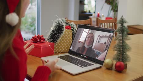 Happy-caucasian-woman-on-video-call-on-laptop-with-female-friend-in-face-mask-at-christmas-time