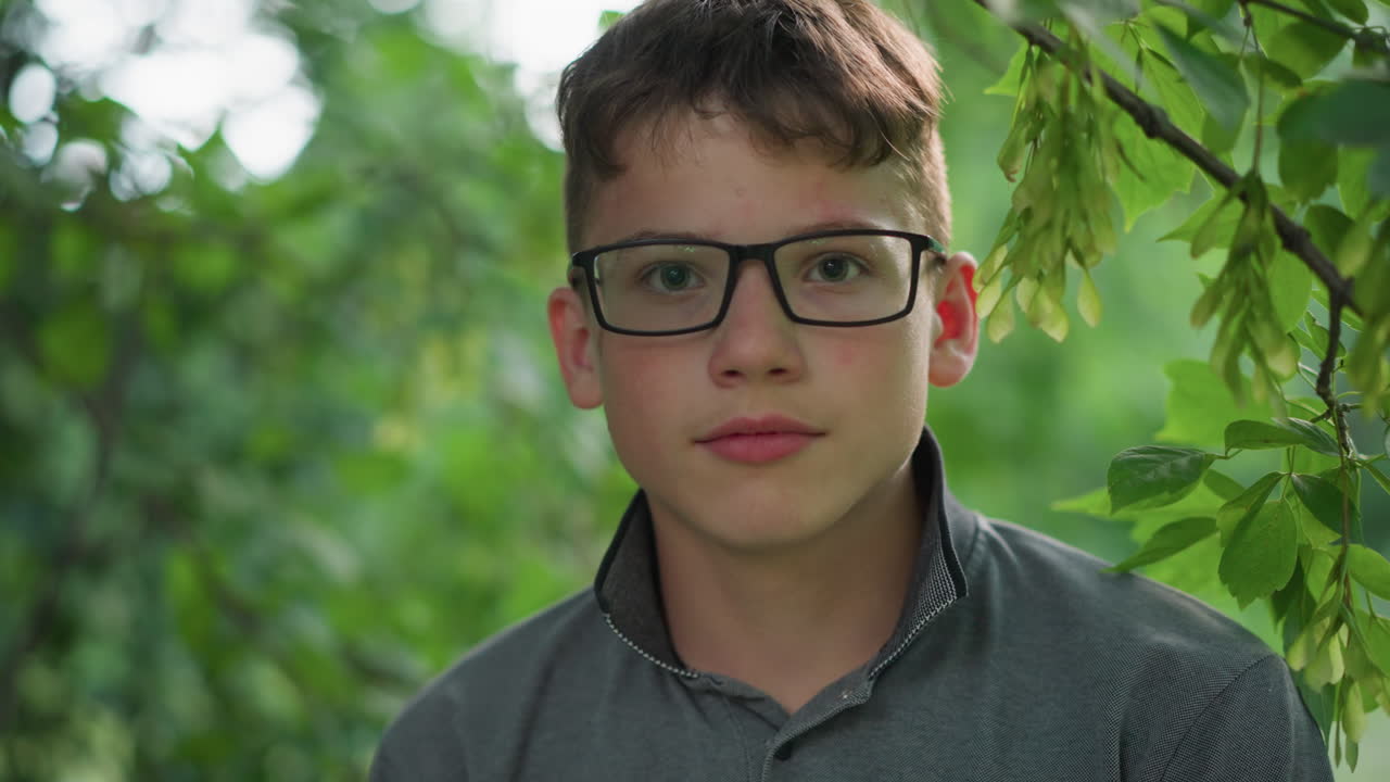 крупный план молодого мальчика с розовыми губами, в очках и сером топе, мигает, глядя прямо в камеру, он окружен пышной зелени с размытыми деревьями на заднем плане