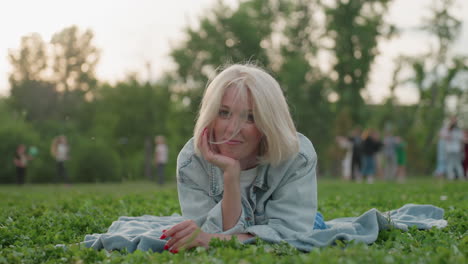 zoom in of young mom lying on picnic blanket supporting head with hand, smiling heartfully, blurred people playing around in park, warm backlight creating relaxed outdoor family vibe