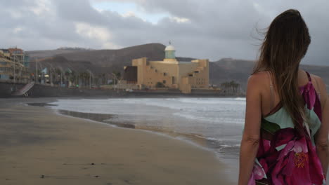 cinematic shot at sunset of a woman walking along las canteras beach and seeing the alfredo kraus auditorium in the background