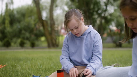 una niña con síndrome de down construyendo una torre de madera sentada en el parque. sus amigos están haciendo artesanías