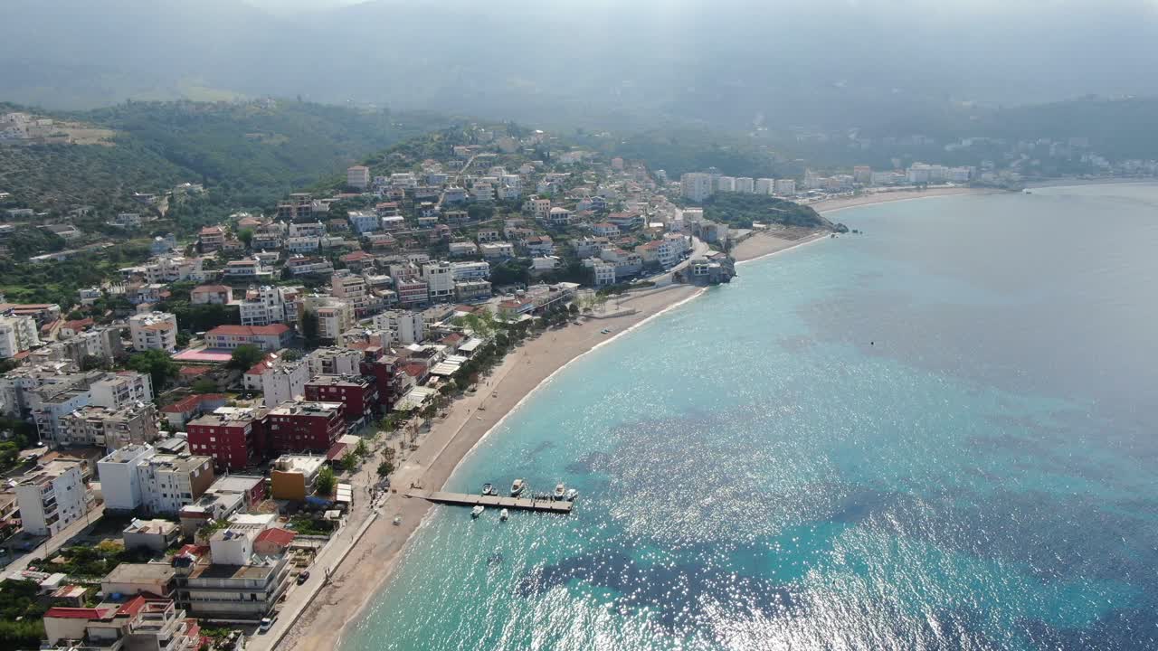 Drone view in Albania flying over a beach with crystal clear blue water ocean, buildings on the harbor and green forest on a sunny day