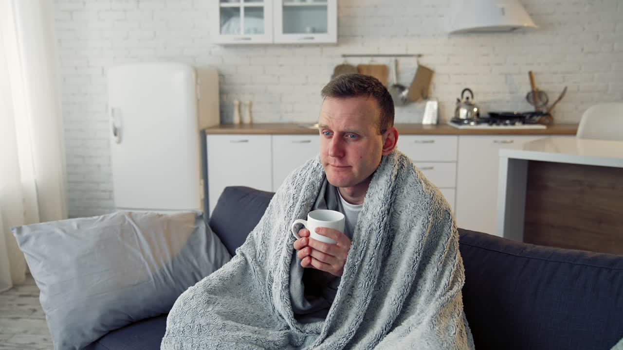 Folk medicine. Sick young man with fever wrapped in warm blanket drinking hot tea. Pandemic.