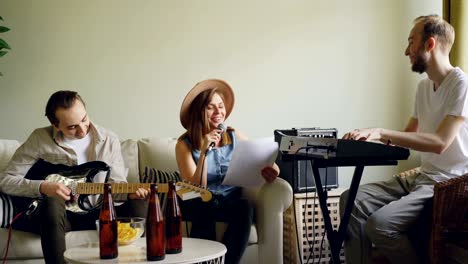 la hermosa vocalista está cantando y riendo durante el ensayo con su banda musical en casa. el tipo barbudo está tocando los teclados y el joven alegre está tocando la guitarra.