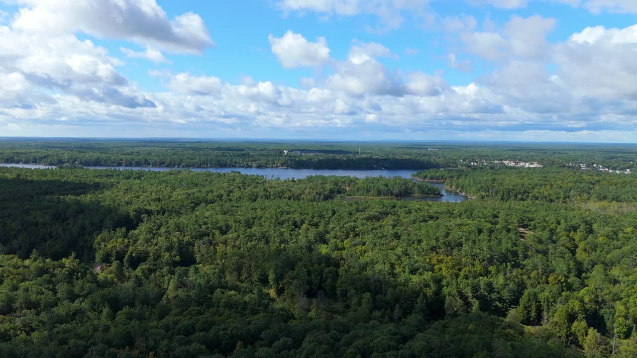 High-altitude drone view over dense Muskoka forest near Gravenhurst, opening toward a long lake under bright clouds and expansive northern landscape