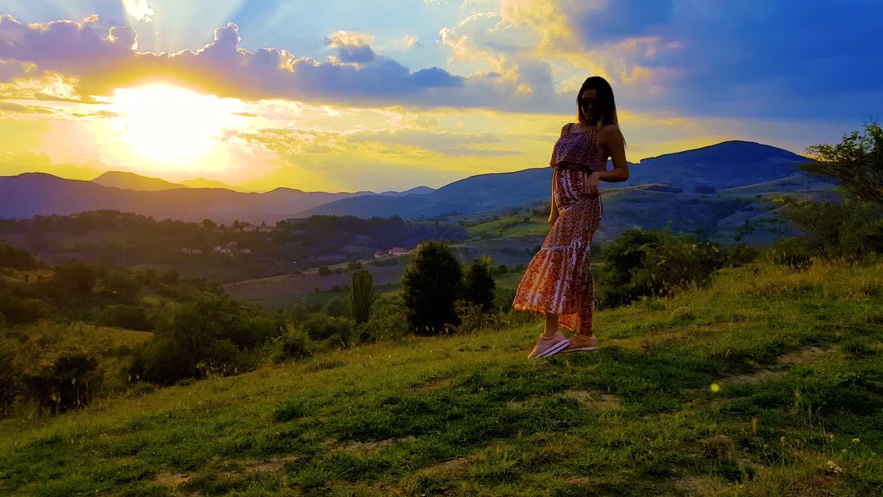puesta de sol sobre las montañas y un cielo nublado con una chica posando, girando y saltando sobre una colina cubierta de hierba