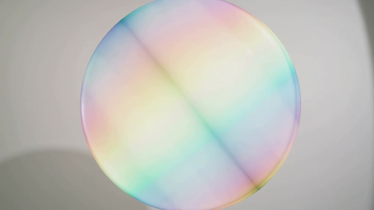 Drifting large soap bubble rotating and shifting thin-film colors under soft light at pale wall