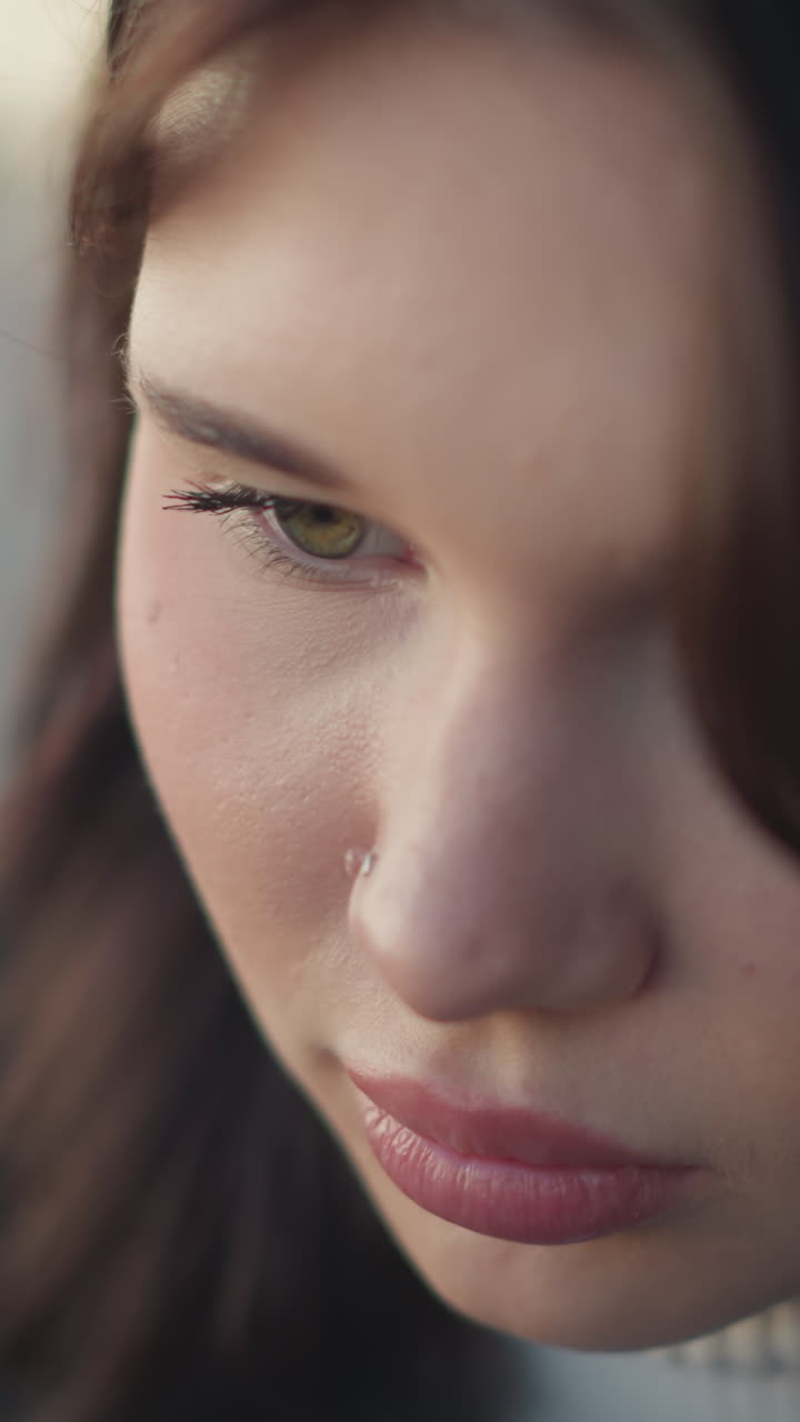 primer plano de una joven con labios rosados y anillo en la nariz, ojos hacia abajo, expresión pensativa, el cabello cae suavemente, con un fondo borroso de colores cálidos