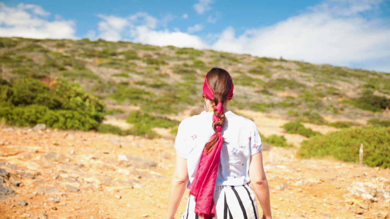 Young brunette with braided fuschia scarf in her hair taking a few steps on a hot sunny day, on a dirt path, on an island, back to camera.