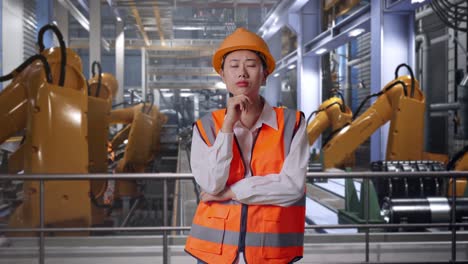asian female engineer with safety helmet thinking and looking around then raising her index finger while standing with industrial robotic arms working