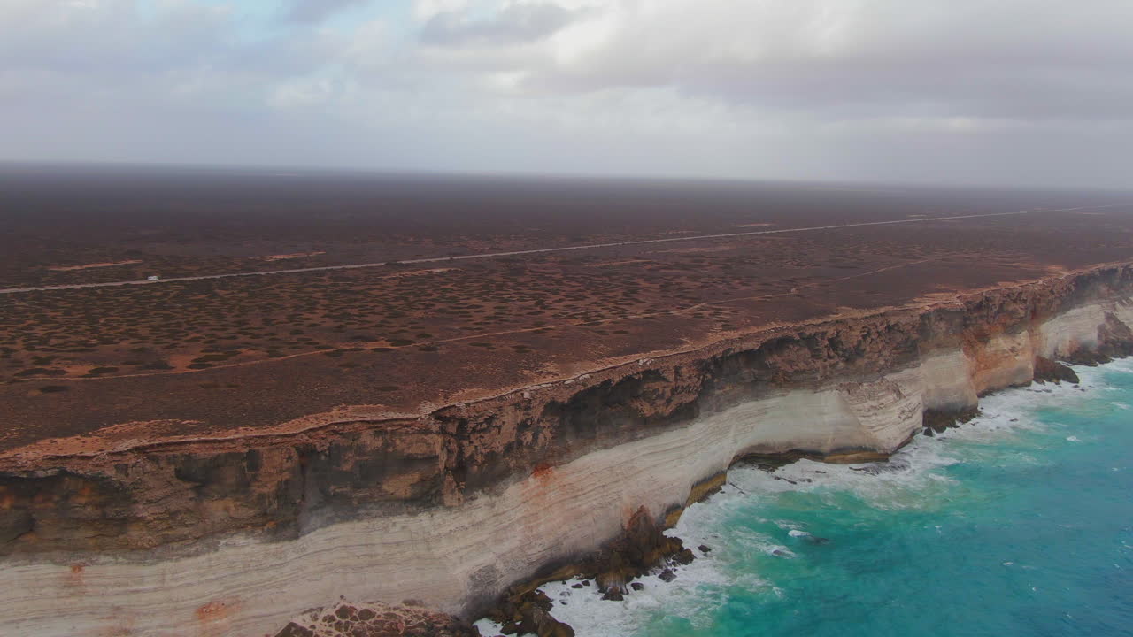 Cinematic drone shot of Campervan on empty road in Nullarbor SA, Australia during cloudy day - Ocean waves crashing against steep rocky coastline