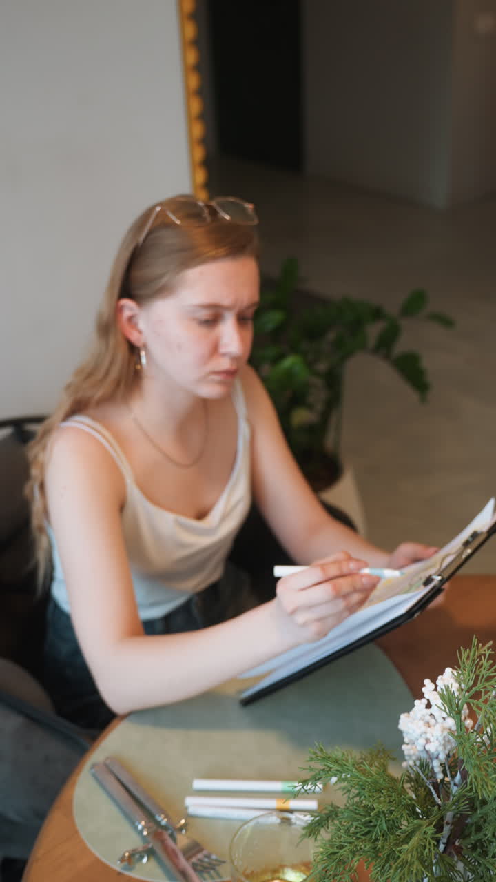 Young woman finalizing checklist at table with markers and clipboard, surrounded by plant and tableware indoor workspace vibe with intent expression suggesting project planning, reservation management