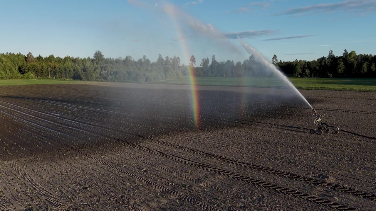 Ripresa al rallentatore con drone di una macchina per l'irrigazione che annaffia un campo, con un arcobaleno visibile nello spruzzo.